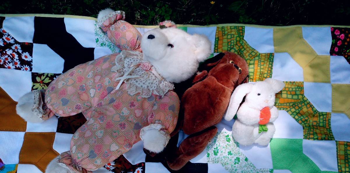 A Teddy Bear, an old stuffed toy dog, and a plush toy bunny on a blanket in the grass.